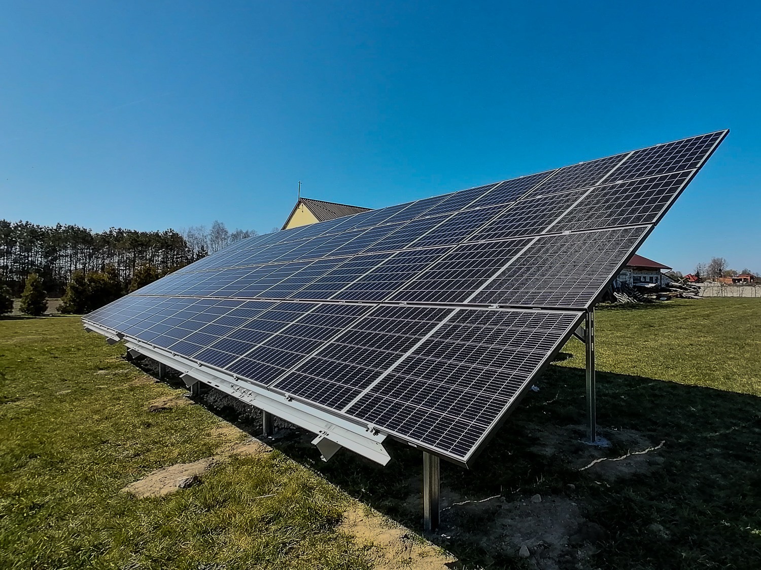 Instalacja paneli słonecznych na trawiastym terenie, z widocznymi elementami konstrukcyjnymi i domem w tle pod błękitnym niebem.