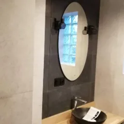 Nowoczesna łazienka z owalnym lustrem podświetlanym LED, czarną umywalką nablatową i baterią w kolorze chromu, drewniany blat i czarne szafki pod umywalką, ściany wyłożone płytkami w odcieniach...