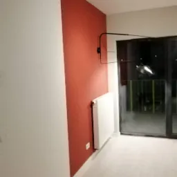 Pomalowany fragment ściany w kolorze terakoty, kontrastujący z bielą pozostałych ścian, z grzejnikiem i lampą ścienną w nowoczesnym wnętrzu po remoncie.