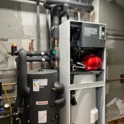 Zestaw pompy ciepła z zasobnikiem wody użytkowej Huch EnTEC, widoczne rury instalacyjne, manometry i elementy sterujące, szara ściana w tle.