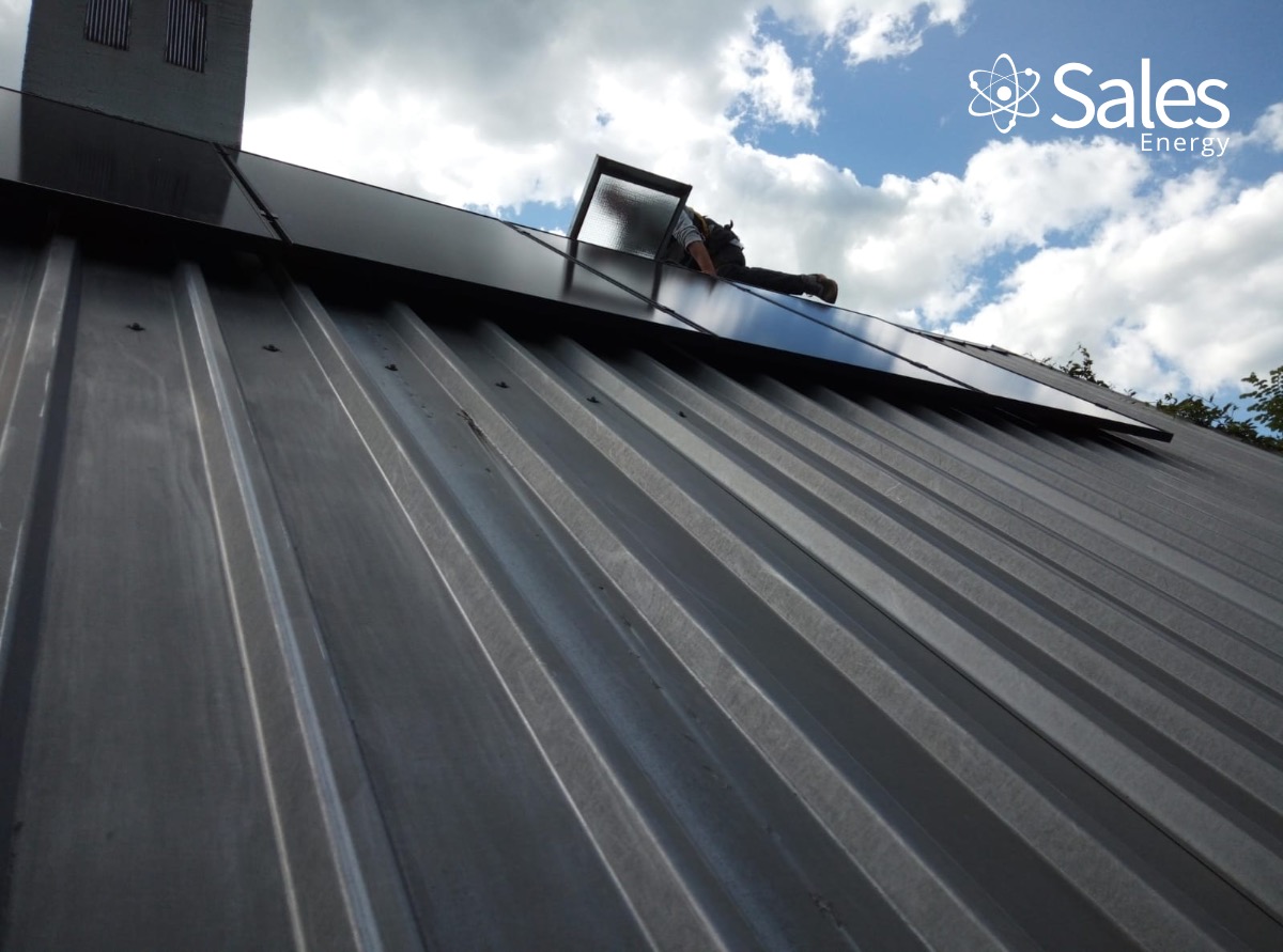 Montaż paneli słonecznych na metalowym dachu, widoczny pracownik w uprzęży i logo firmy Sales Energy na tle pochmurnego nieba.