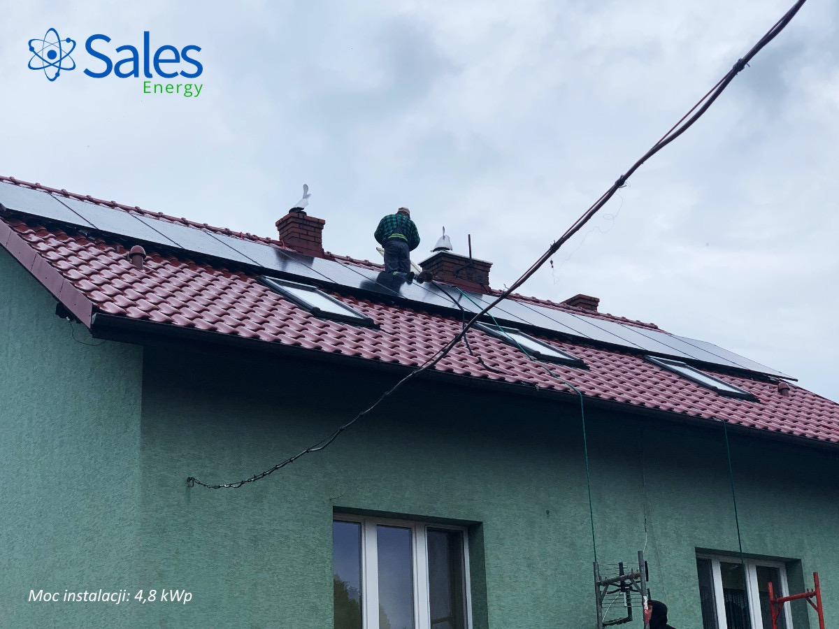 Montaż paneli słonecznych na dachu domu z czerwoną dachówką, widoczny pracownik w zielonej kurtce i logo firmy Sales Energy w lewym górnym rogu.