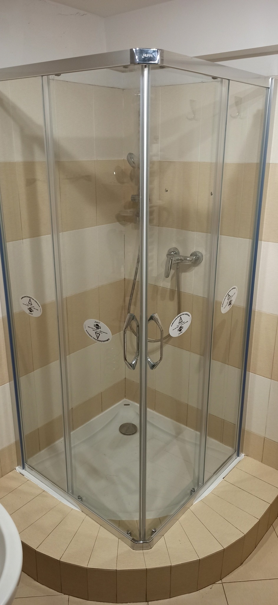 Narożna kabina prysznicowa z chromowanymi profilami i szklanymi drzwiami, widoczne naklejki ostrzegawcze na szybach, zamontowana bateria i deszczownica, beżowe płytki na ścianach i podłodze.