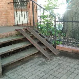 Zewnętrzne schody z rampami dla wózków inwalidzkich, wykonane z metalu i umieszczone przy ceglanym budynku z ozdobną kratą w oknie, widoczne pozostałości po myciu.