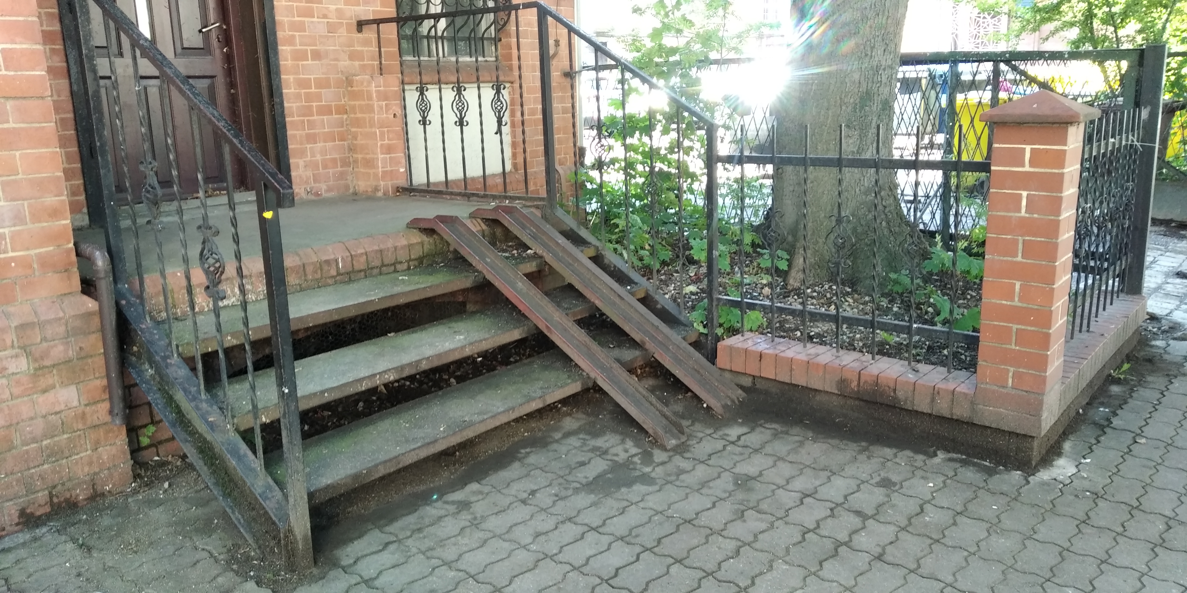 Zewnętrzne schody z rampami dla wózków inwalidzkich, wykonane z metalu i umieszczone przy ceglanym budynku z ozdobną kratą w oknie, widoczne pozostałości po myciu.