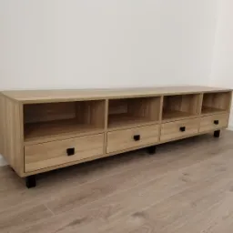 Jasna szafka RTV z drewna dębowego z czterema otwartymi półkami i trzema szufladami z czarnymi, kwadratowymi uchwytami, na niskich, czarnych nóżkach.