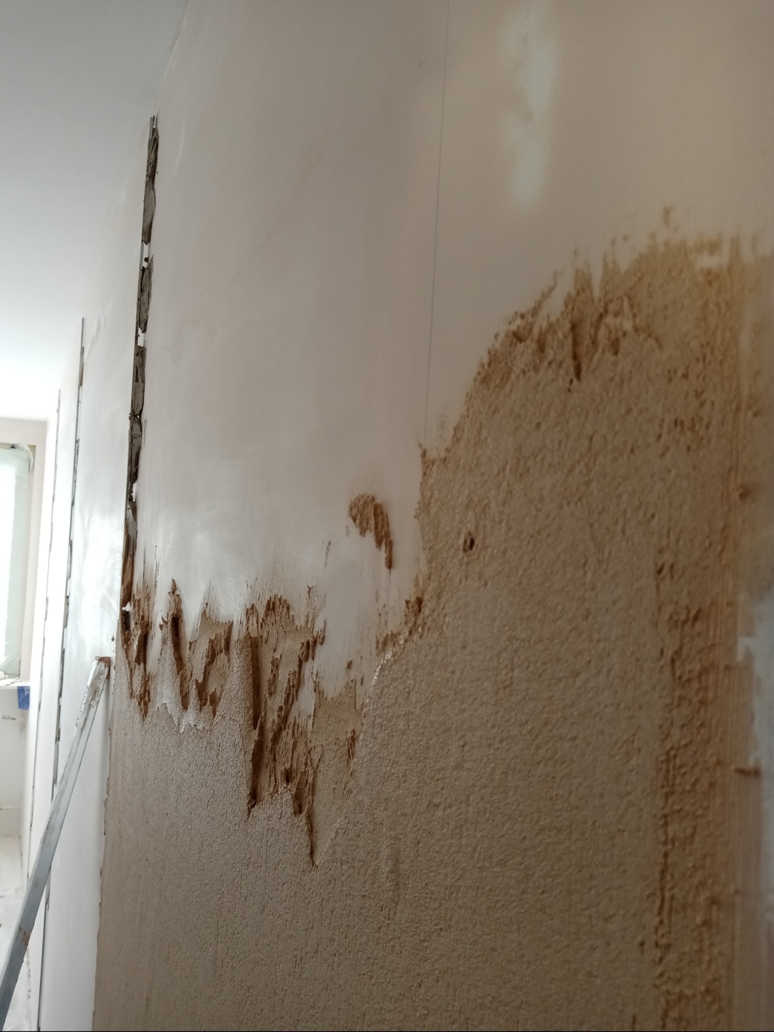 Ściana w trakcie nakładania gładzi, widoczne fragmenty surowej ściany i nałożona warstwa gładzi w kolorze beżowym, listwa przypodłogowa w lewym dolnym rogu.