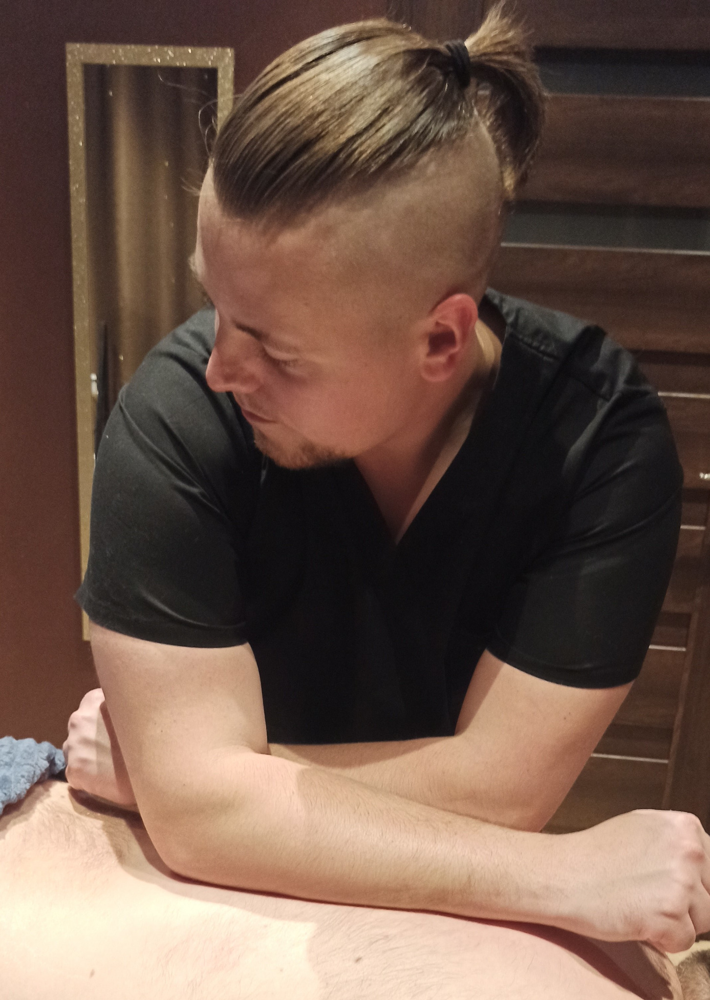 Mężczyzna w czarnym uniformie wykonuje masaż pleców w Chorzowie, używając łokci. W tle widoczny element dekoracyjny ściany.