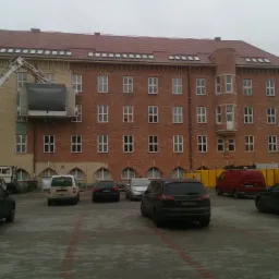 Elewacja budynku z cegły częściowo umyta, z użyciem podnośnika koszowego, na parkingu zaparkowane samochody.