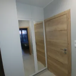 Szafa wnękowa z lustrzanymi drzwiami i drewnianym frontem, widoczny fragment pokoju z łóżkiem w odbiciu lustra, minimalistyczny design.