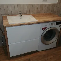 Biała umywalka nablatowa z drewnianym blatem, wbudowana w szafkę z pralką LG w łazience z beżowymi płytkami.