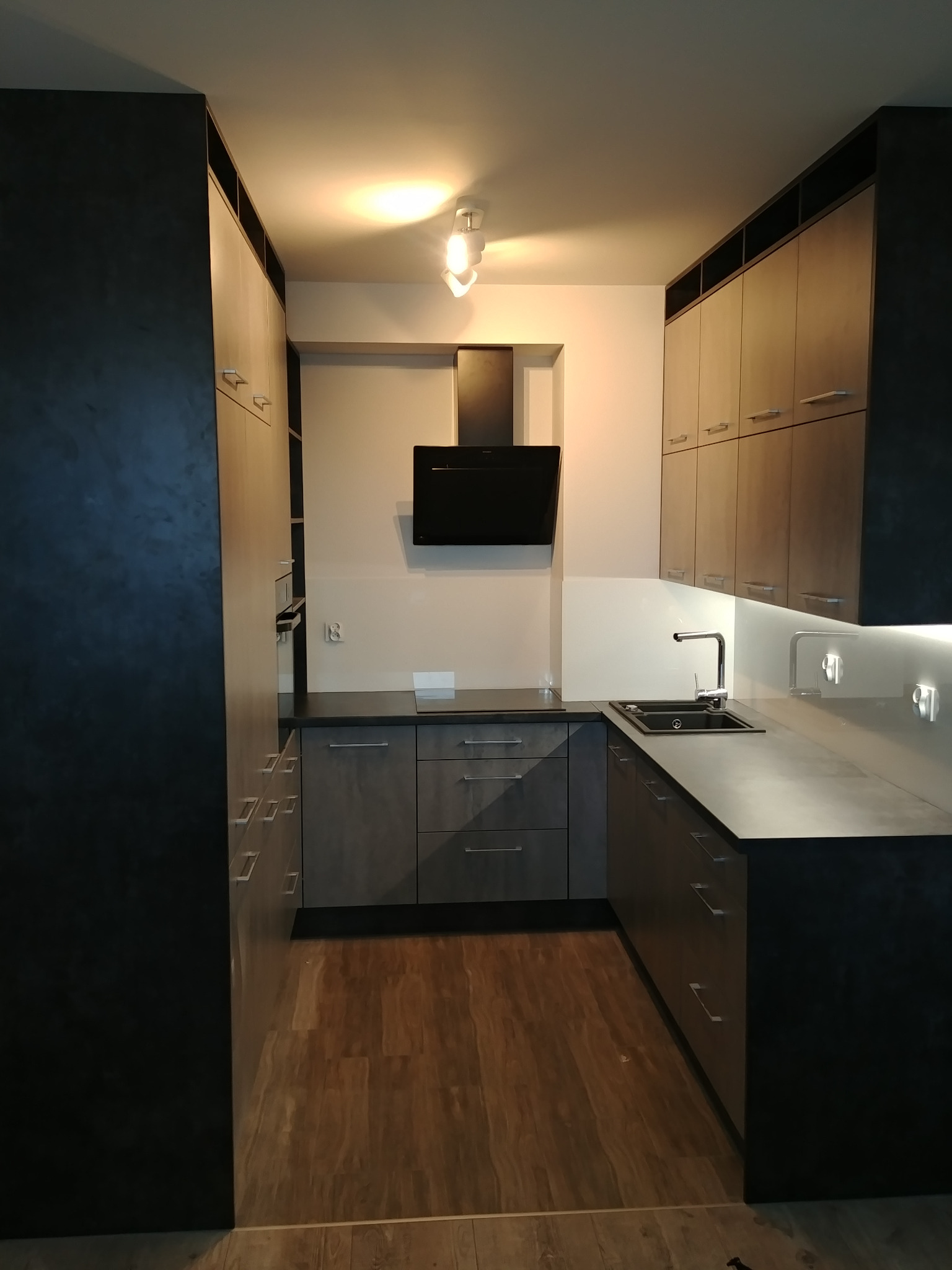 Nowoczesna kuchnia z szarymi frontami szafek, czarnym okapem i blatem, oświetleniem podszafkowym oraz drewnianą podłogą.