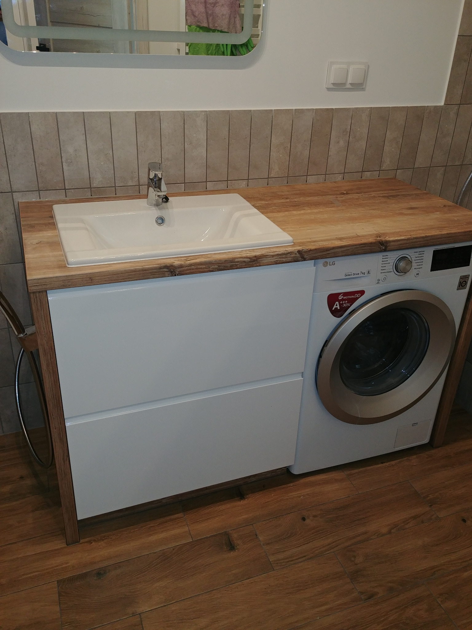 Biała umywalka nablatowa z drewnianym blatem, wbudowana w szafkę z pralką LG w łazience z beżowymi płytkami.