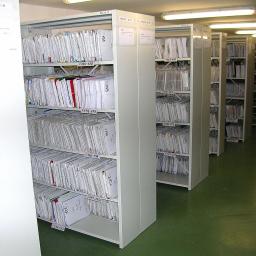 Usługi Archiwalne Jarosław Żymańczyk Ewa Żymańczyk - Rzędy metalowych regałów wypełnionych segregatorami z dokumentami w archiwum, widoczne etykiety z numeracją na półkach i segregatorach, zielona podłoga.