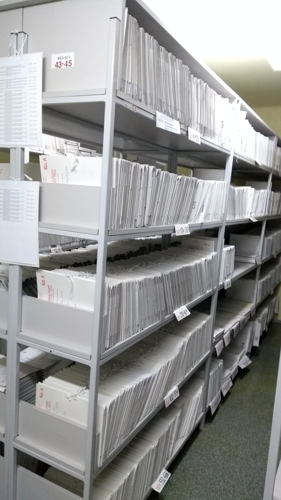 Metalowe regały wypełnione segregatorami z dokumentami, oznaczone etykietami z numerami, widoczne w pomieszczeniu archiwum.