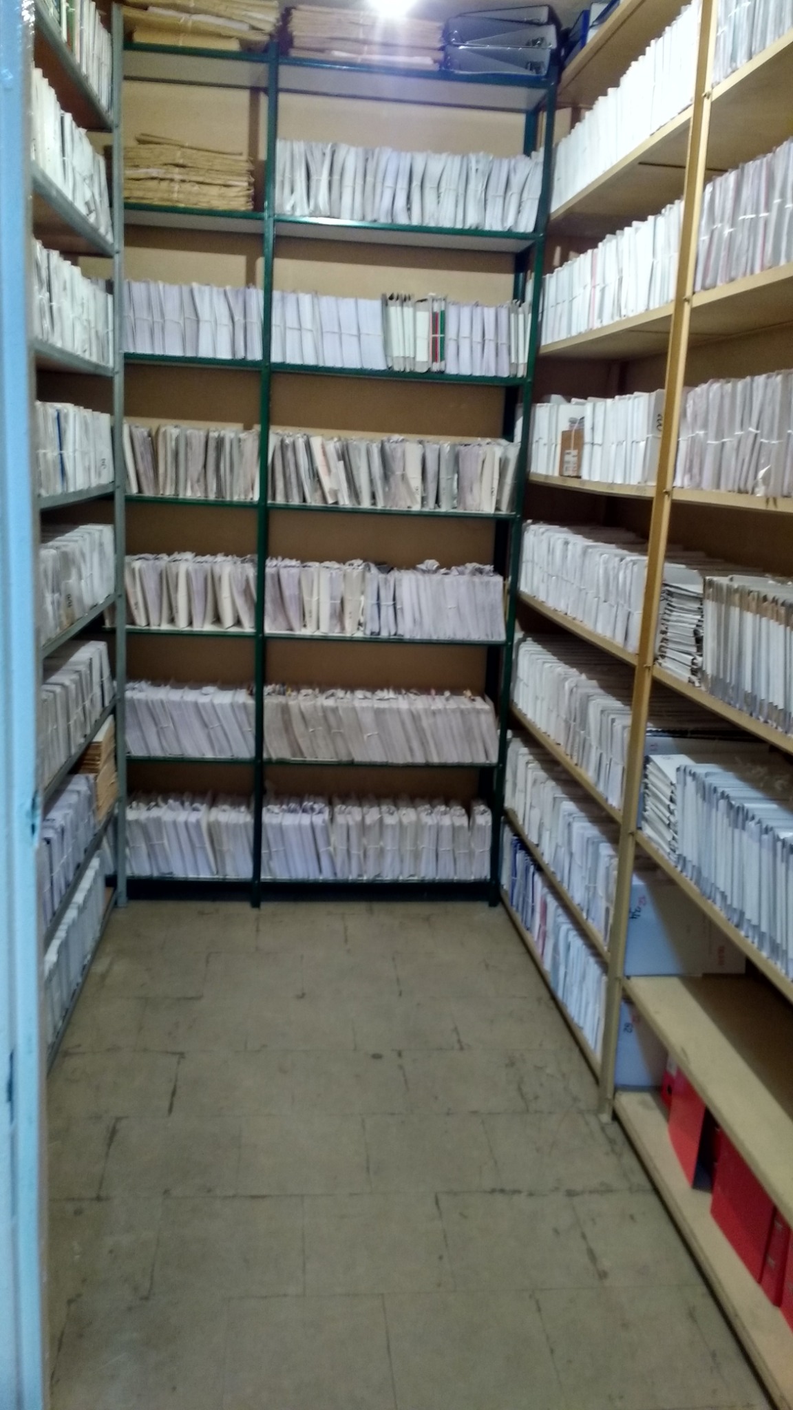 Pełne regały z dokumentami w archiwum, widok z perspektywy korytarza, segregatory ułożone w rzędach.