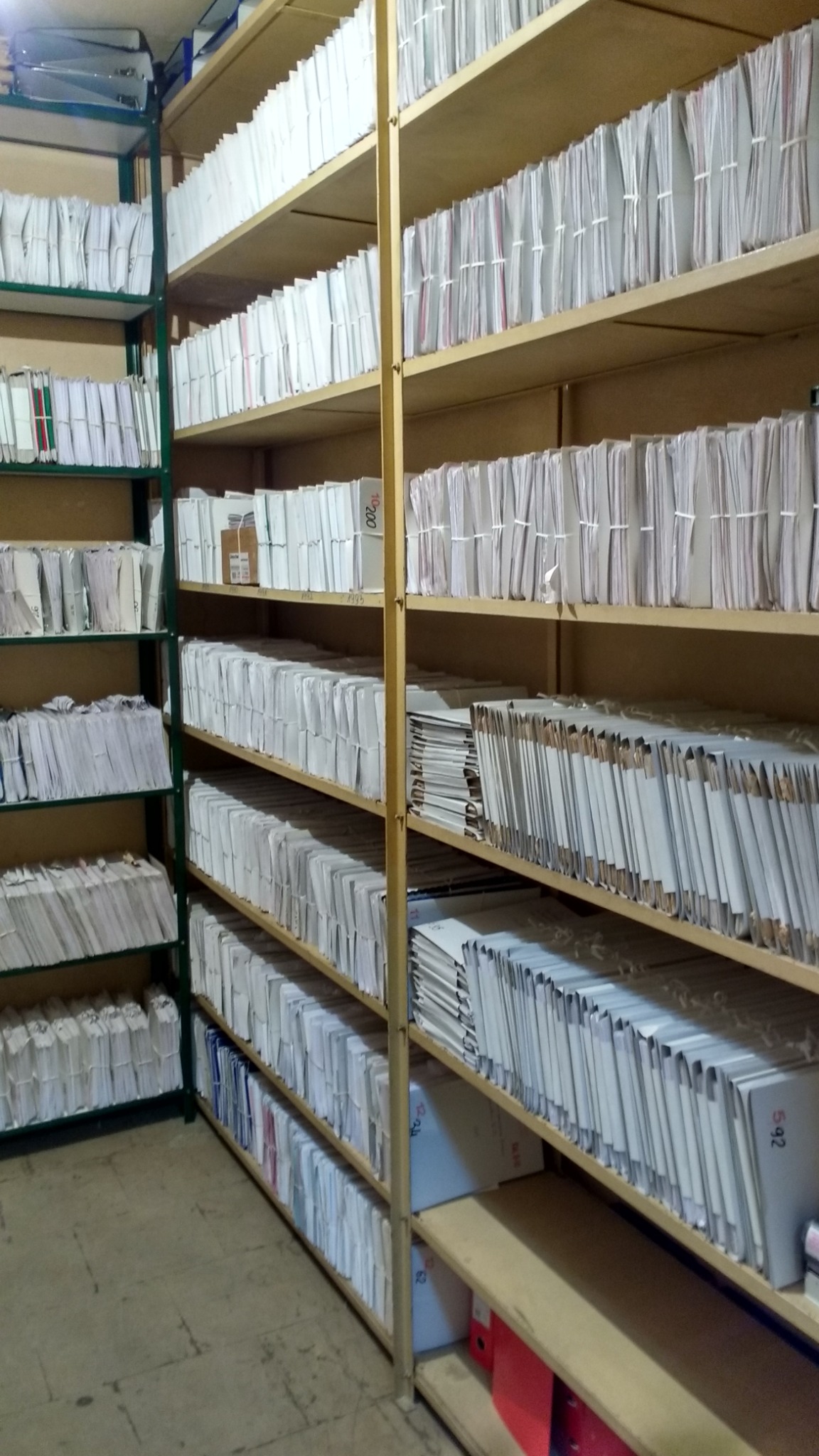 Półki regału wypełnione segregatorami i teczkami z dokumentami, widoczne różne systemy archiwizacji i organizacji dokumentów.