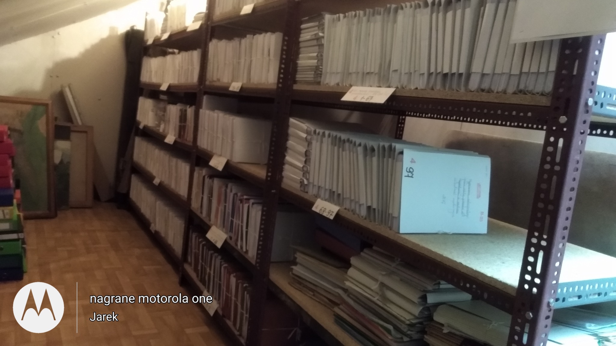 Półki regału wypełnione segregatorami i pudłami z dokumentami w pomieszczeniu na poddaszu.