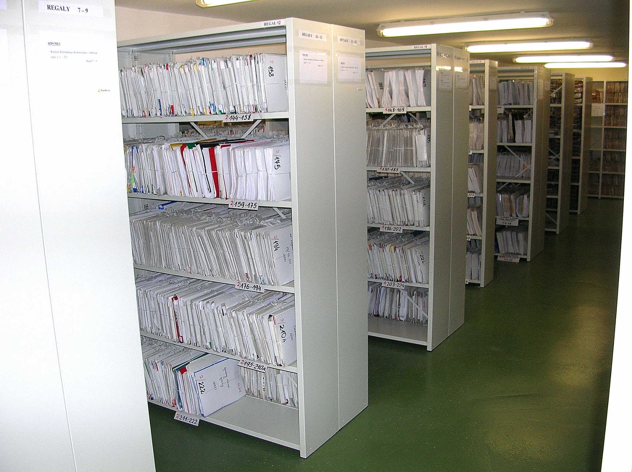 Rzędy metalowych regałów wypełnionych segregatorami z dokumentami w archiwum, widoczne etykiety z numeracją na półkach i segregatorach, zielona podłoga.