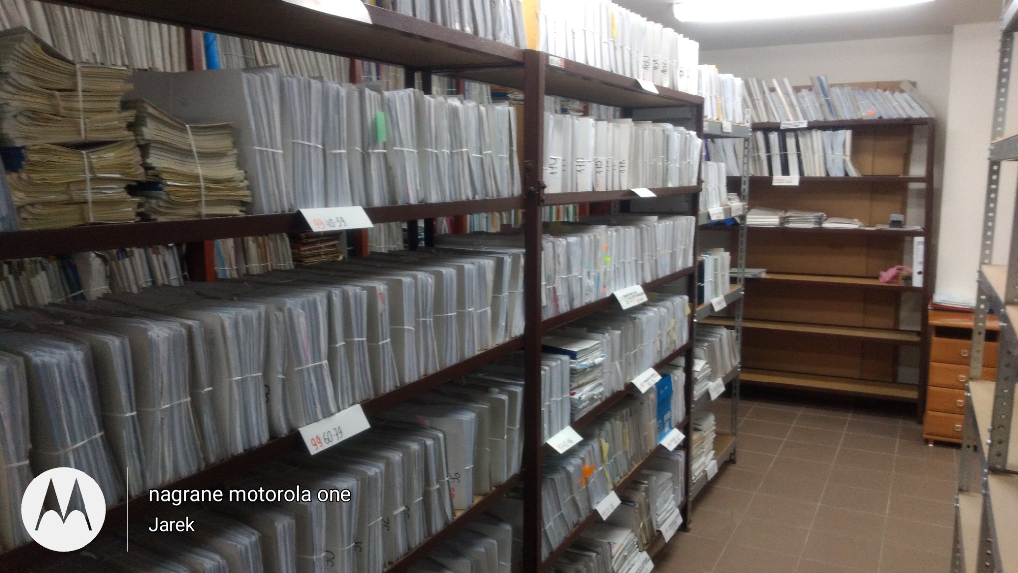 Regały magazynowe wypełnione segregatorami i dokumentami, z etykietami identyfikacyjnymi, tworząc system archiwizacji w pomieszczeniu z brązowymi płytkami podłogowymi.