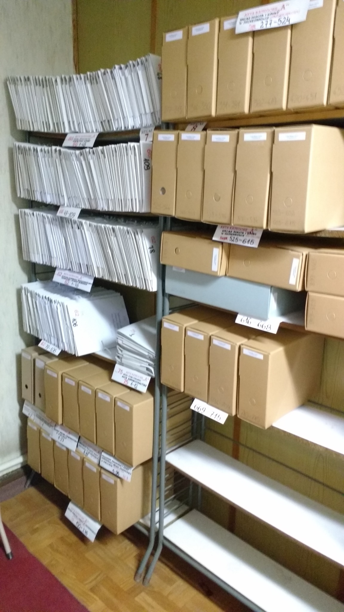 Półki regału wypełnione segregatorami i kartonami z dokumentami, oznaczonymi etykietami z numerami, w archiwum biurowym.