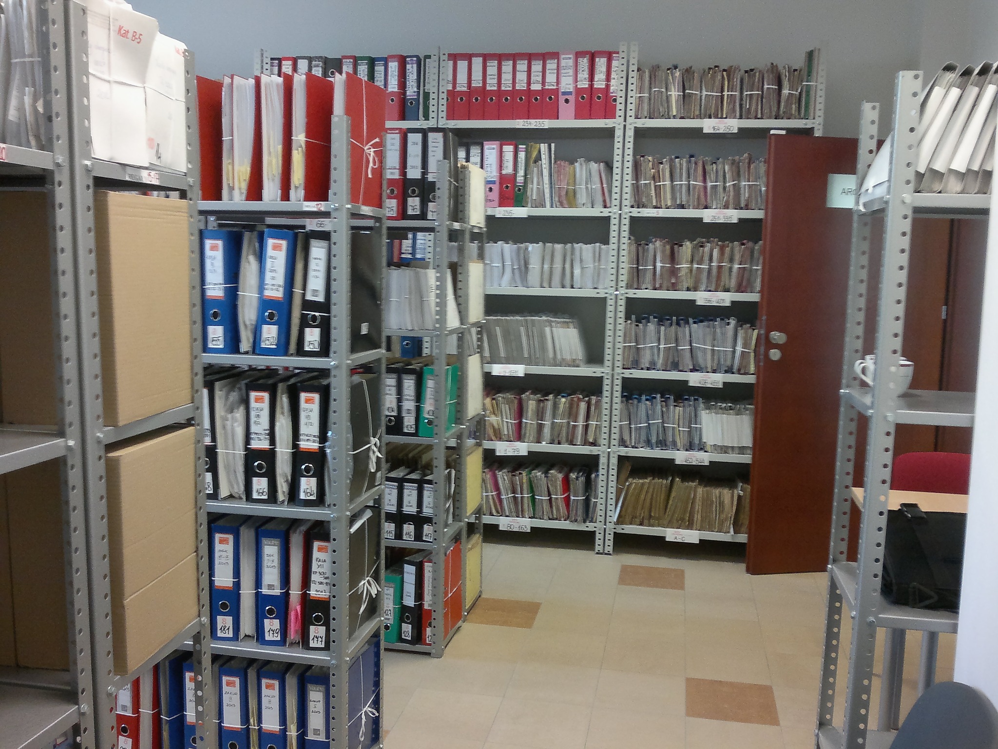 Rzędy metalowych regałów wypełnione segregatorami i dokumentami w archiwum, widoczne oznaczenia na segregatorach i półkach.