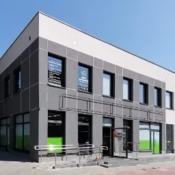 Nowoczesny budynek biurowy w odcieniach szarości z dużymi oknami, na których widoczne są plakaty informacyjne, w tym logo i dane kontaktowe firmy.