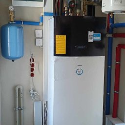 Soltimus Sp. z o.o. - Biała pompa ciepła Daikin z podłączonymi rurami w kolorze niebieskim i czerwonym, z żółtą naklejką ostrzegawczą i naklejką z kodem QR, obok niebieski zbiornik wyrównawczy i skrzynka elektryczna...