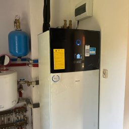 Soltimus Sp. z o.o. - Biała pompa ciepła Daikin w narożniku pomieszczenia, z widocznymi elementami instalacji hydraulicznymi i elektrycznymi, w tym niebieski zbiornik wyrównawczy i rury doprowadzające, z naklejkami...