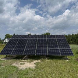 Soltimus Sp. z o.o. - Panel fotowoltaiczny na łące, z widocznym stosem drewna i domem w tle. Błękitne niebo z chmurami. Instalacja na gruncie, widoczne podpory.