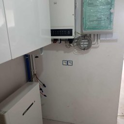 Soltimus Sp. z o.o. - Biała instalacja fotowoltaiczna z inwerterem i skrzynką elektryczną na ścianie. Widoczne kable i przewody. Elementy instalacji częściowo zasłonięte przez szafkę.