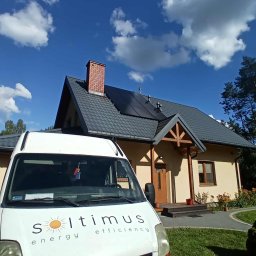 Soltimus Sp. z o.o. - Dom z panelami słonecznymi na dachu, widziany zza firmowego samochodu Soltimus. Błękitne niebo z chmurami w tle. Widoczny komin z cegły i zadbany ogród.