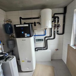Soltimus Sp. z o.o. - Zmontowana instalacja pompy ciepła Daikin w pomieszczeniu z białymi płytkami. Widoczne zbiorniki, rury, zawory i skrzynka elektryczna. Surowy betonowy sufit.