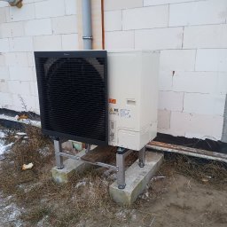 Soltimus Sp. z o.o. - Zewnętrzna jednostka pompy ciepła Daikin, zamontowana na betonowych podstawach przy ścianie budynku z białego bloczka. Widoczne rury i resztki śniegu na ziemi.