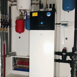 Soltimus Sp. z o.o. - Biała pompa ciepła Daikin w kotłowni z instalacją grzewczą, zbiornikami i rozdzielaczem ogrzewania podłogowego. Widoczne rury i zawory.