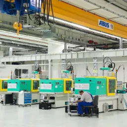 Wnętrze hali produkcyjnej z rzędem wtryskarek Arburg w kolorach zielonym i żółtym, obsługiwanych przez pracownika w niebieskim kombinezonie, widoczny suwnica ABUS.