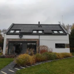 Nowoczesny dom jednorodzinny z panelami fotowoltaicznymi na dachu, widoczne okna dachowe, antena satelitarna i dekoracyjne trawy wzdłuż ścieżki.