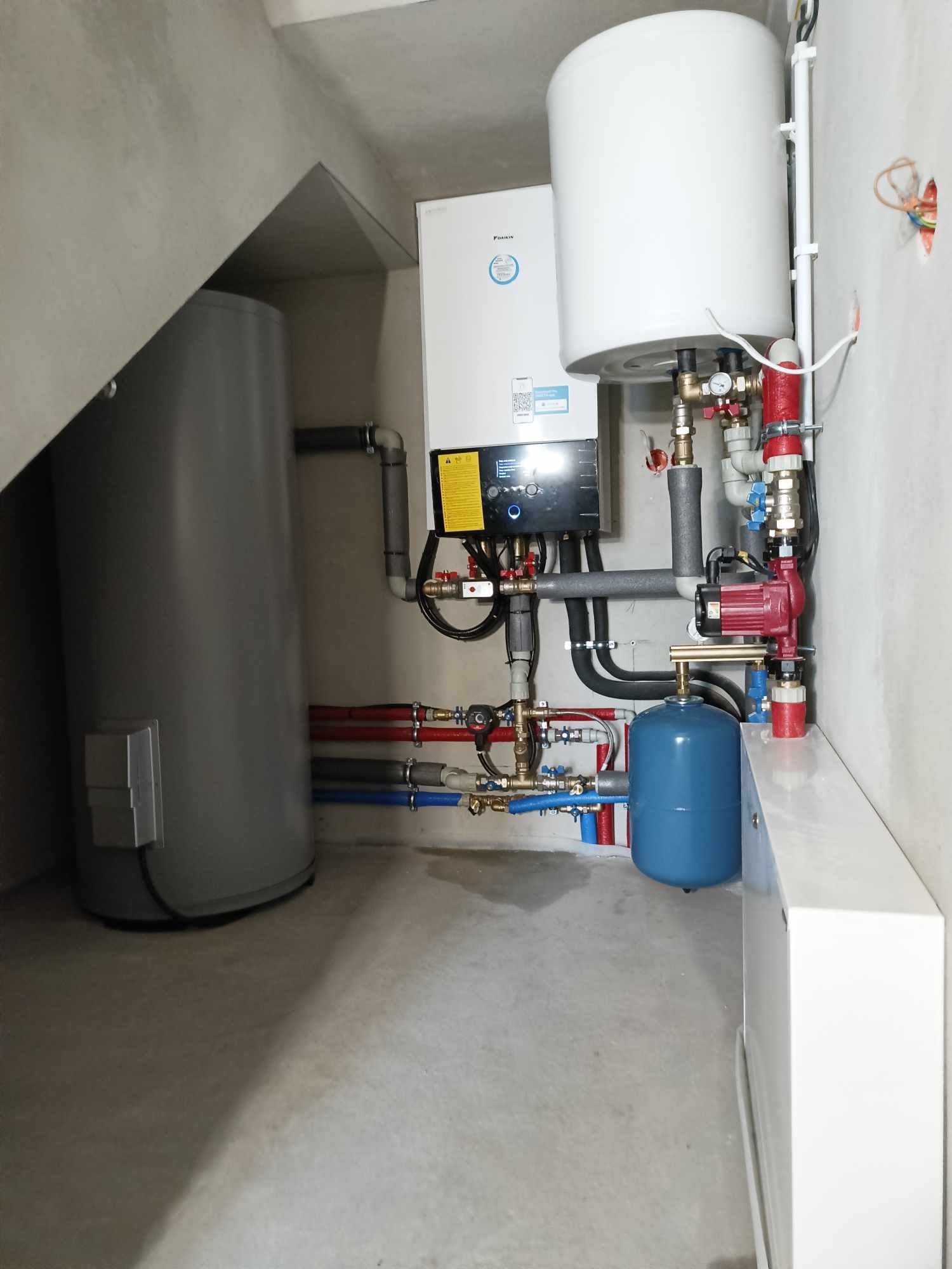 Zestaw urządzeń grzewczych w kotłowni: zasobnik ciepłej wody użytkowej, pompa obiegowa, naczynie przeponowe i kocioł kondensacyjny Daikin, połączone rurami w kolorach czerwonym i niebieskim.