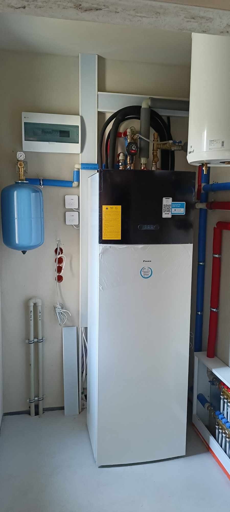 Biała pompa ciepła Daikin z czarnym panelem górnym, podłączona do instalacji z rurami w kolorze niebieskim i czerwonym, z widocznym zbiornikiem wyrównawczym i skrzynką elektryczną na ścianie.