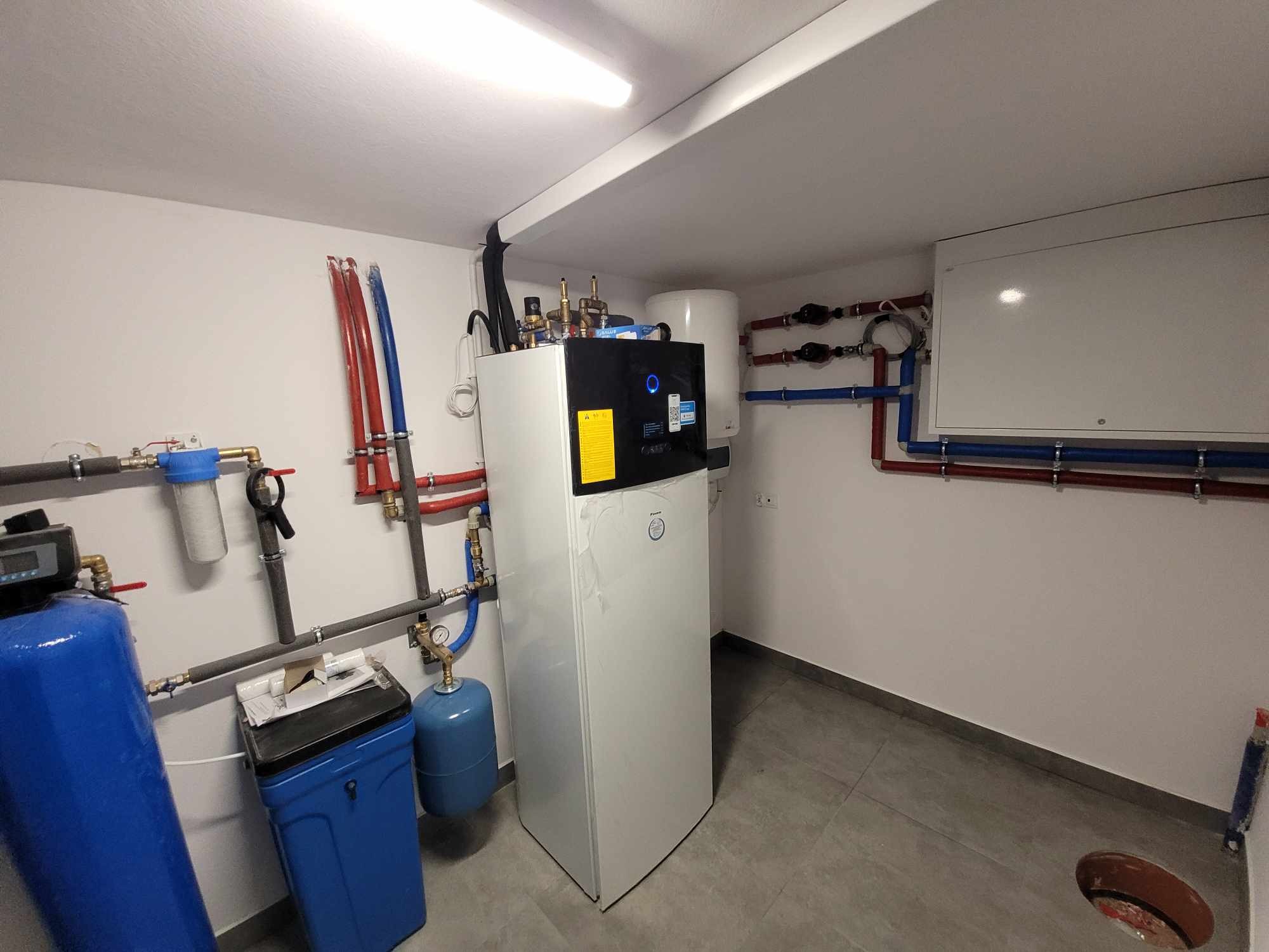 Wnętrze kotłowni z zainstalowaną białą pompą ciepła Daikin, zbiornikiem buforowym, systemem filtrów wody i rozbudowaną instalacją hydrauliczną z rur miedzianych i plastikowych.