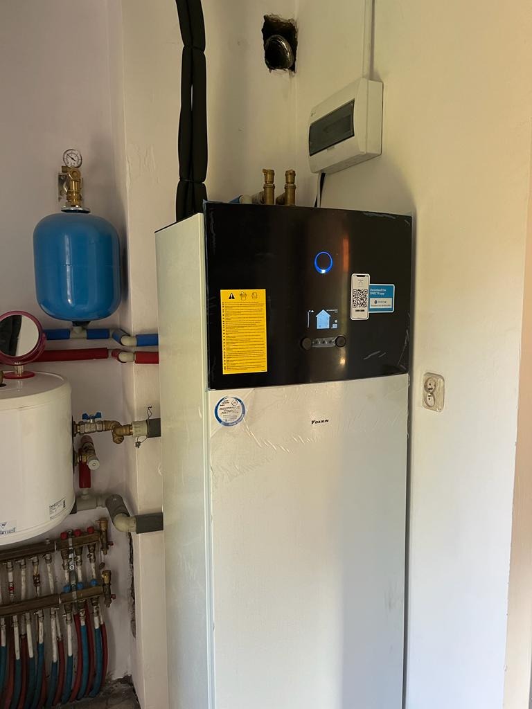 Biała pompa ciepła Daikin w narożniku pomieszczenia, z widocznymi elementami instalacji hydraulicznymi i elektrycznymi, w tym niebieski zbiornik wyrównawczy i rury doprowadzające, z naklejkami...