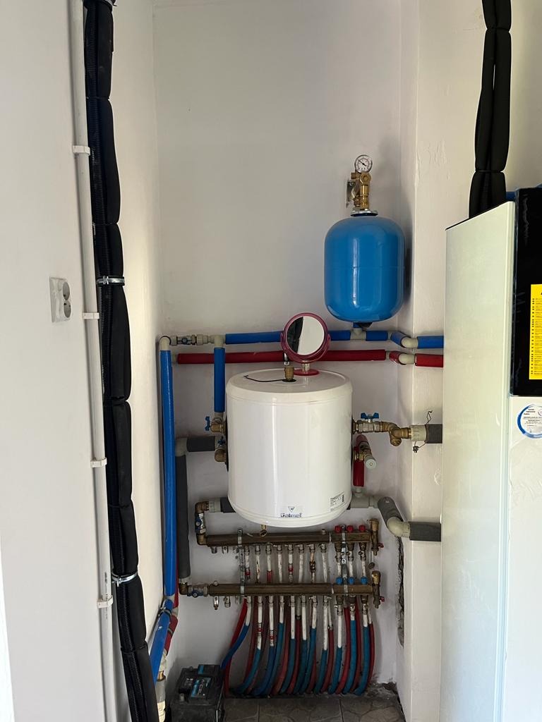 Węzeł cieplny w kotłowni: biały zbiornik buforowy Galmet, niebieskie naczynie przeponowe z manometrem, rozdzielacz z licznymi zaworami i pętlami rur w kolorach czerwonym i niebieskim, czarne rury...