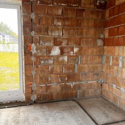 Mały Elektryk - Instalacja elektryczna w surowym wnętrzu: kable w peszlach prowadzone po ścianie z cegły, widoczne puszki elektryczne. Okno z widokiem na zieleń w tle.