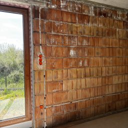 Mały Elektryk - Ściana z pustaków z instalacją elektryczną w trakcie montażu, widoczne puszki i przewody. Okno z widokiem na zieleń w tle. Surowy stan wykończenia.