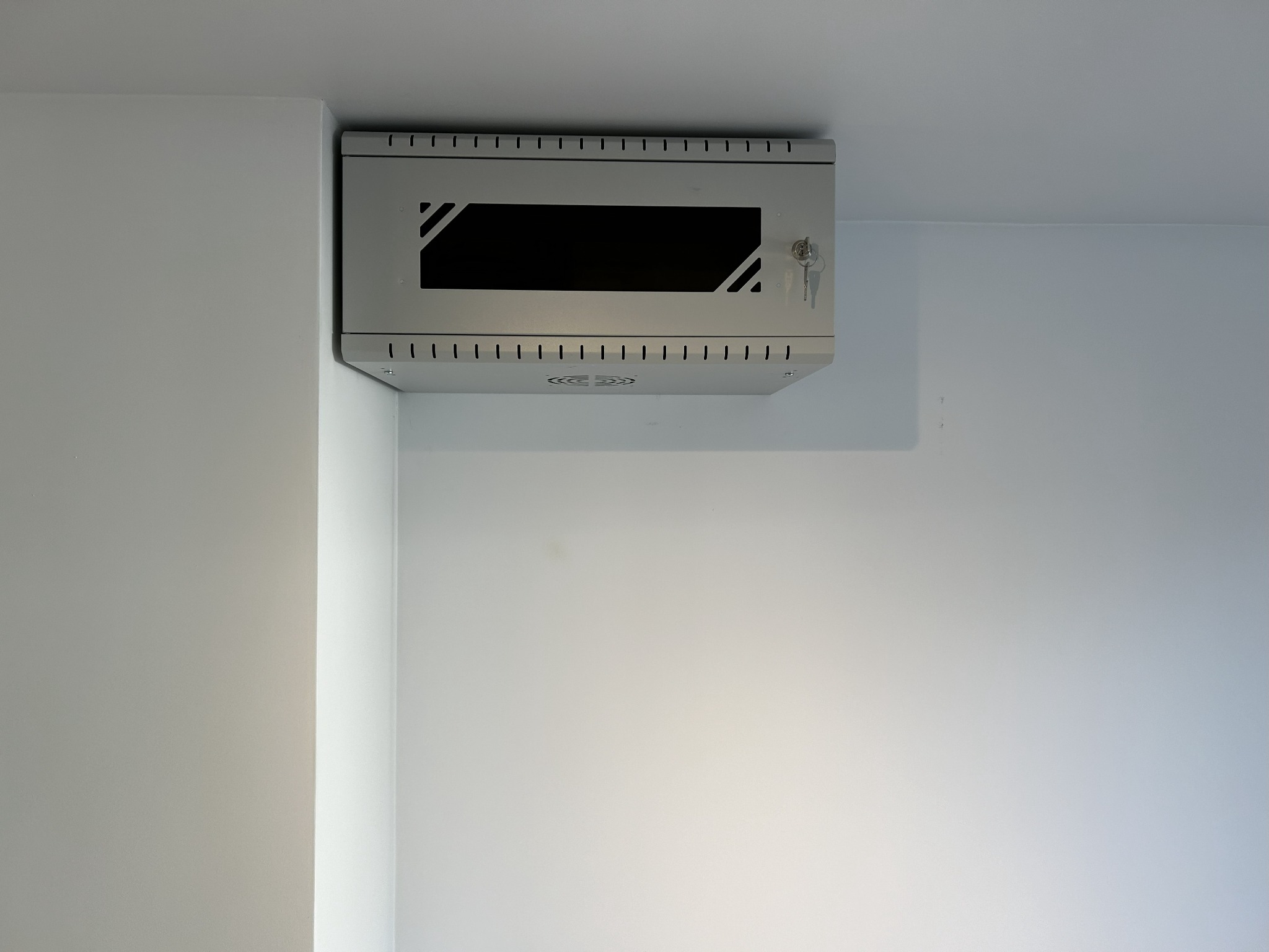 Szara szafka naścienna z wentylacją i kluczem w zamku, zamontowana na białej ścianie. Widoczne otwory wentylacyjne i zamek z kluczem.