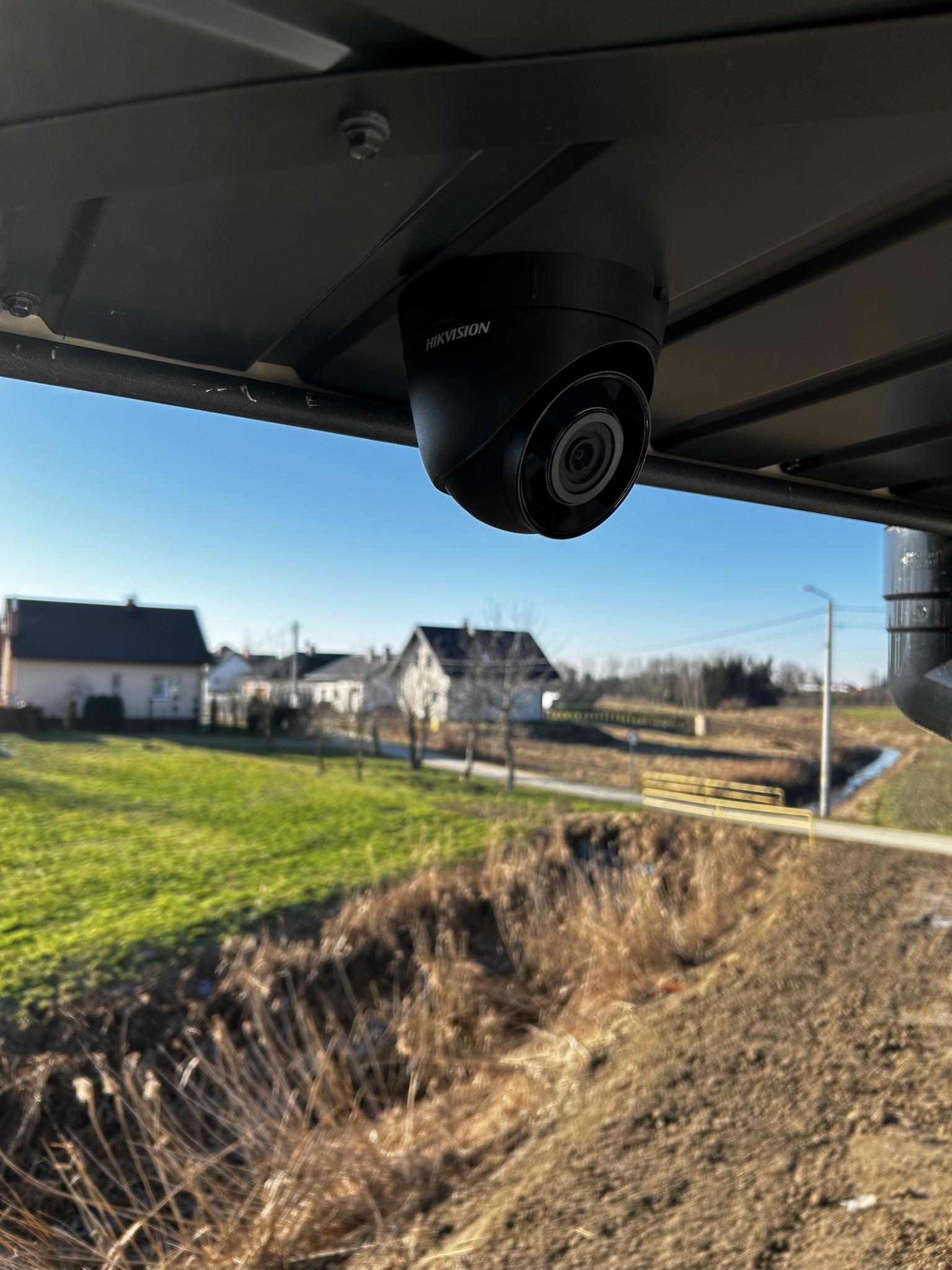 Kamera Hikvision zamontowana na zewnątrz, z widokiem na domy i krajobraz. Czarny korpus kamery kontrastuje z jasnym niebem w tle.