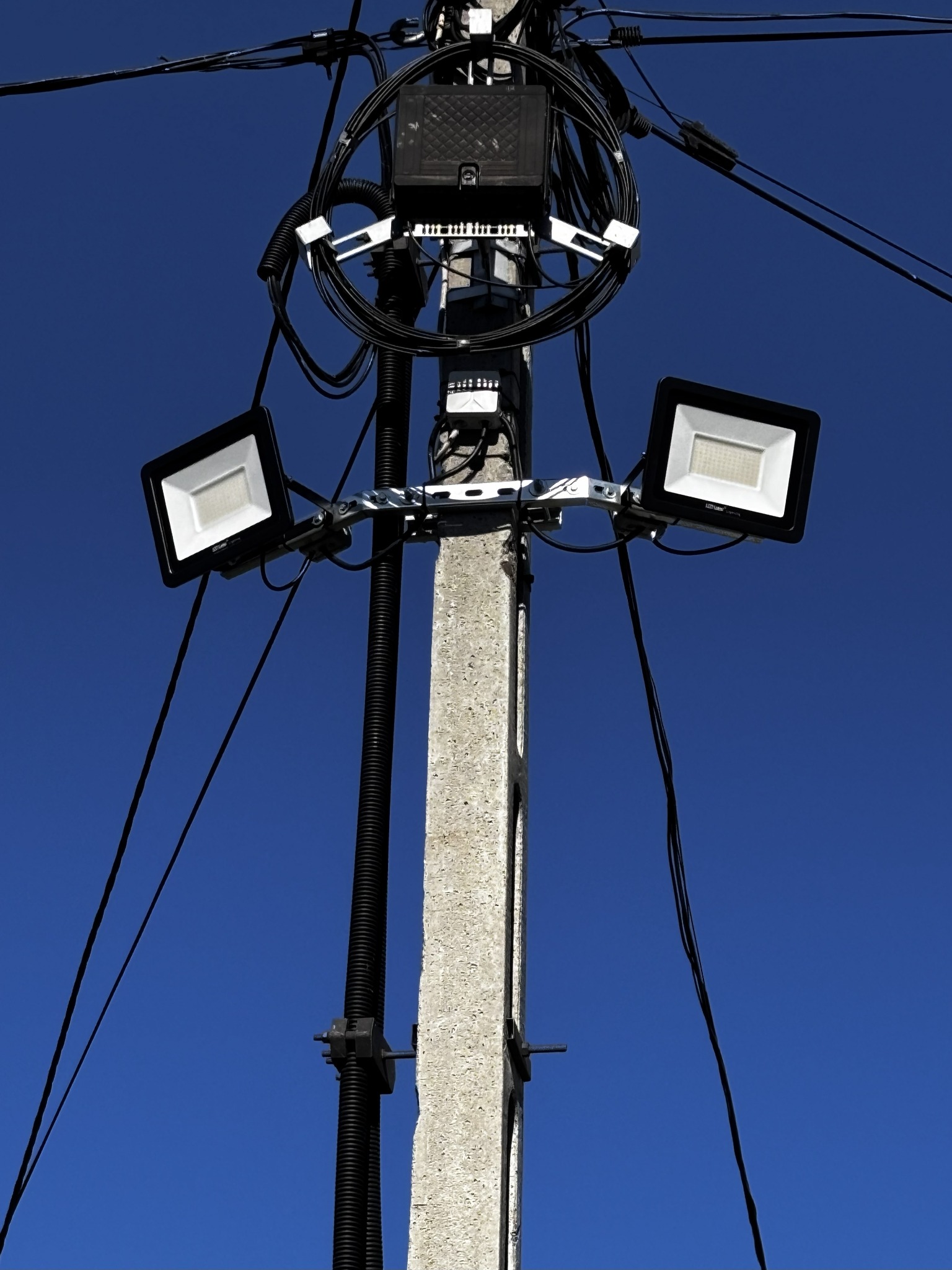Betonowy słup z zamontowanymi dwoma reflektorami LED i okablowaniem na tle intensywnego, błękitnego nieba. Widoczne elementy instalacji elektrycznej.