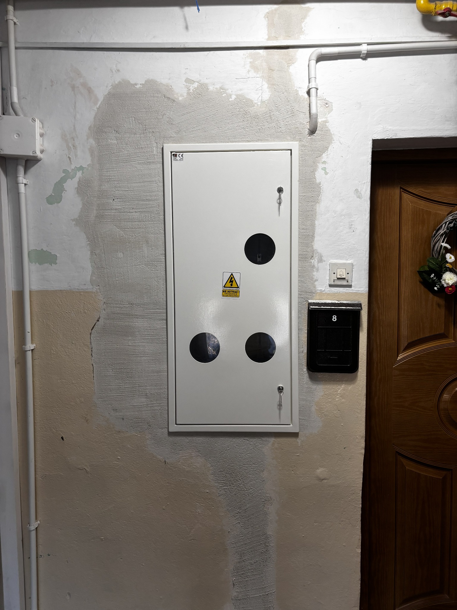 Biała skrzynka elektryczna z trzema okrągłymi otworami i symbolem ostrzegawczym, zamontowana na nierównej ścianie obok skrzynki na listy i drewnianych drzwi.