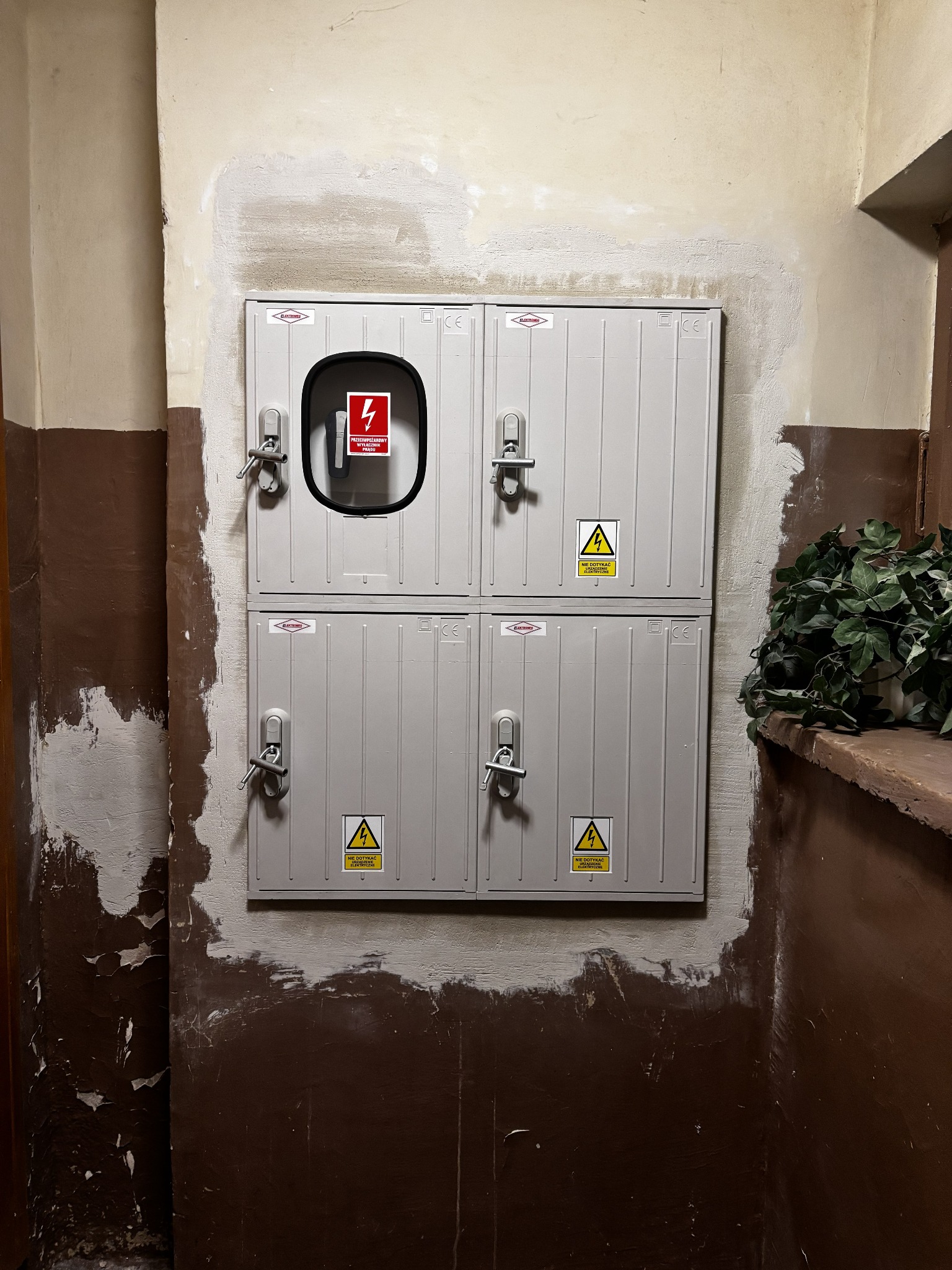 Szara, czteroczęściowa skrzynka elektryczna z ostrzeżeniami, zamontowana na ścianie w trakcie remontu. Widoczne ślady po malowaniu i rośliny doniczkowe obok.
