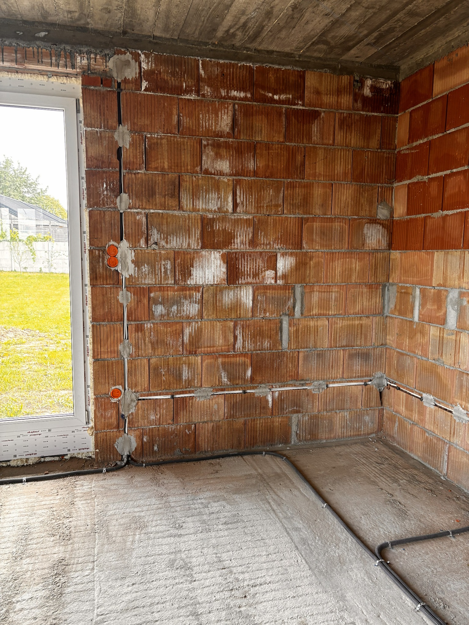 Instalacja elektryczna w surowym wnętrzu: kable w peszlach prowadzone po ścianie z cegły, widoczne puszki elektryczne. Okno z widokiem na zieleń w tle.